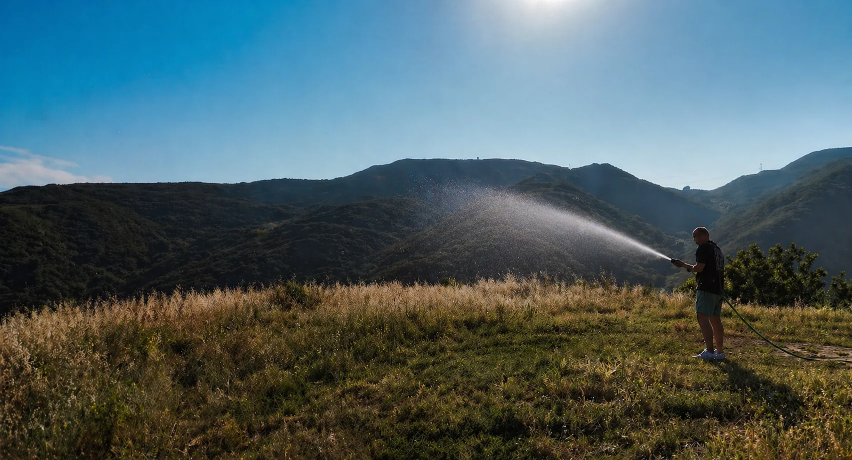 A person holding a garden hose, water spraying a short, low arc that barely reaches the field in front